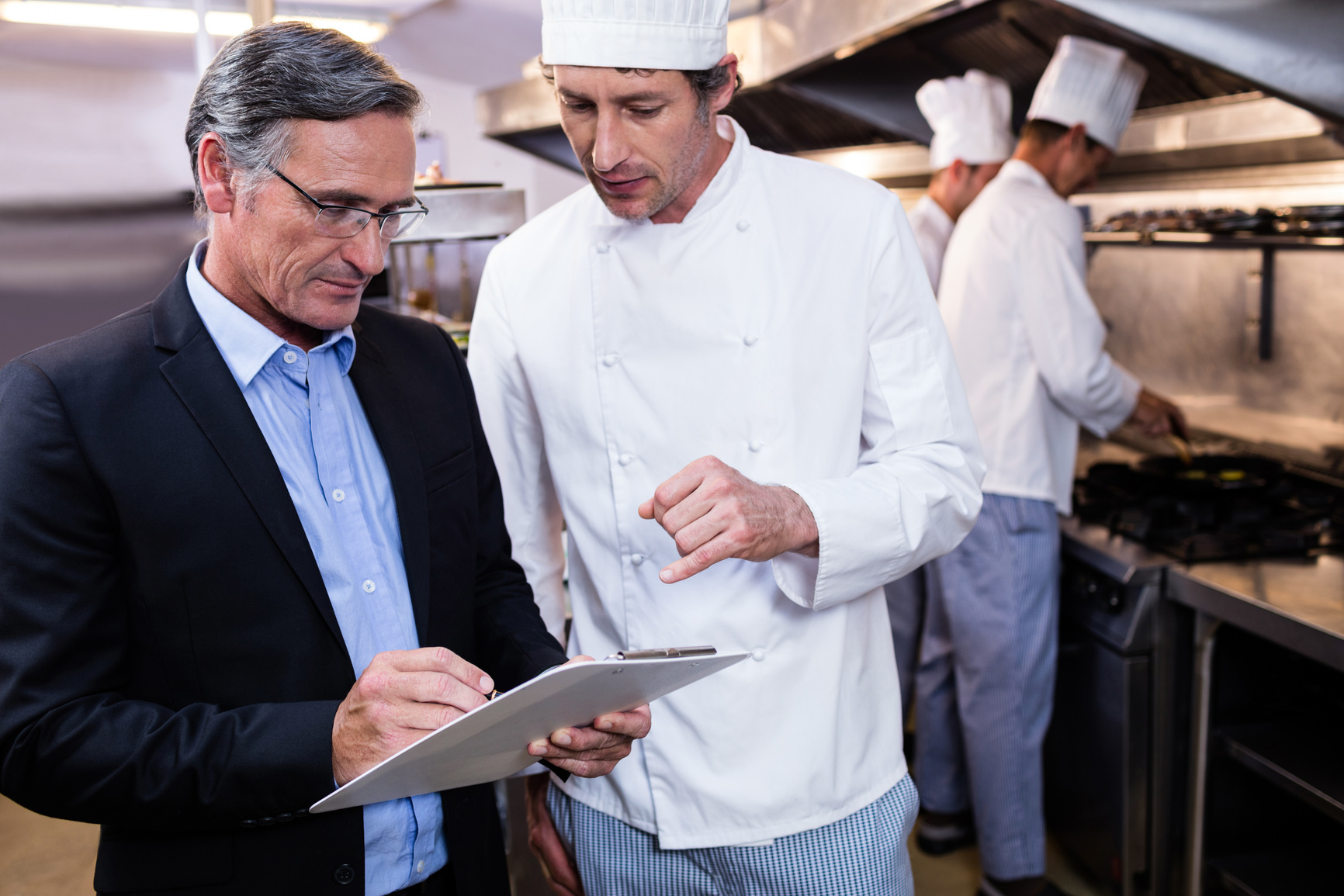 Male restaurant manager writing on clipboard while interacting to head chef